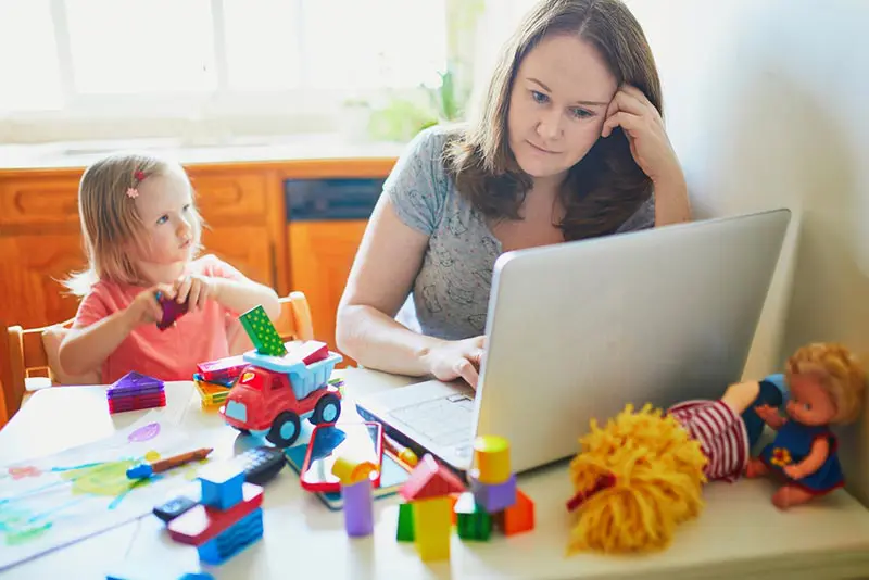 White mom working on laptop at the table with young child sitting next to her with toys all over table.