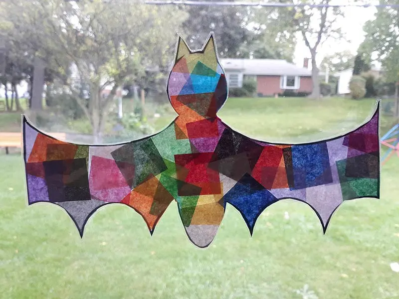 Halloween bat shaped paper suncatcher hanging in window looking into grassy backyard.