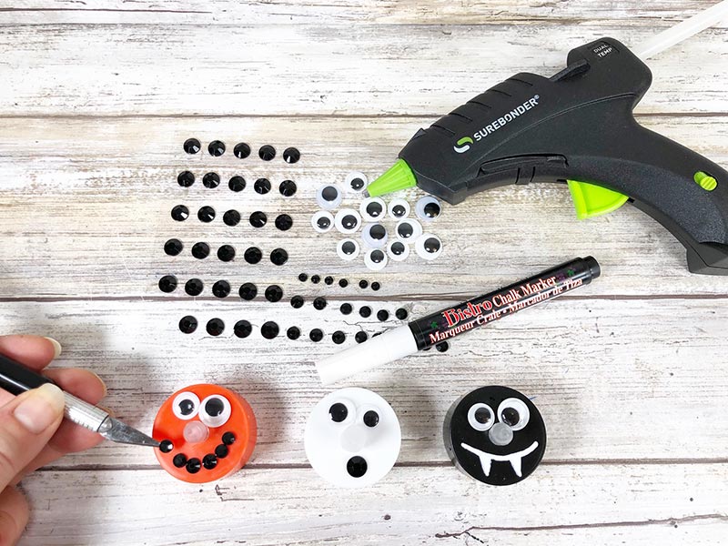 Woman's hand holding craft knife to place black rhinestones on orange tea light. Orange, white, and black tea lights next to each other decorated to look like faces. Black rhinestones, chalk marker, and hot glue gun lay nearby.