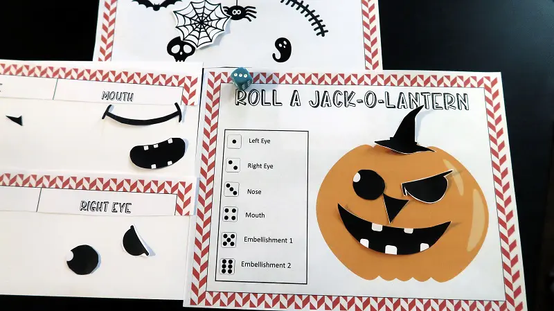Roll a Jack-O-Lantern dice game printables on black table with blue die. Cut out eyes, nose and mouth piece arranged on pumpkin to make a face.