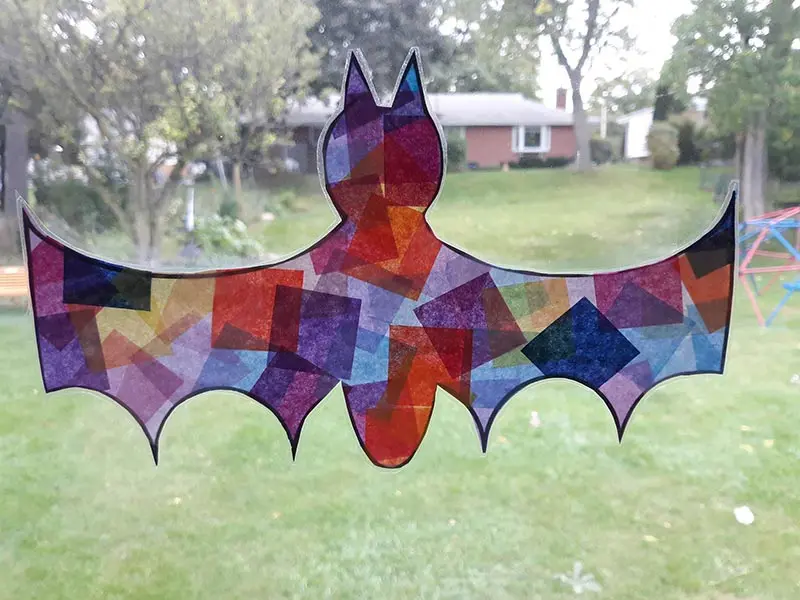 Colorful tissue paper makes bat look like faux stained glass suncatcher in window with grassy background.