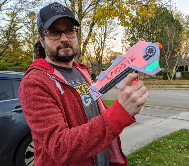 Adult white man with dark hair and beard wearing a hat, glasses, and red hoodie holding a Laser X Evolution blaster lit up for red team and full hit points.