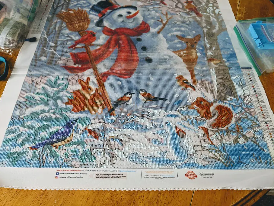 Happy snowman winter scene diamond art painting kit from Diamond Art Club on table with some rhinestone pieces applied along the bottom third of the canvas.
