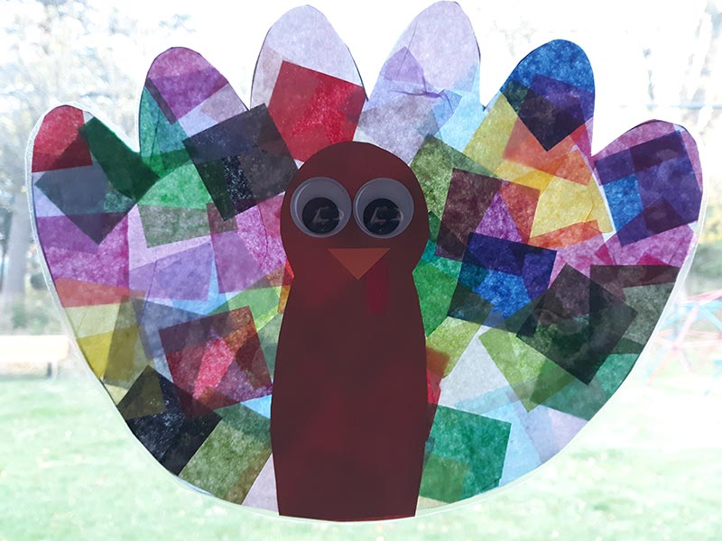 Turkey craft with tissue paper feathers and construction paper body hanging in window with sun shining through the feathers.