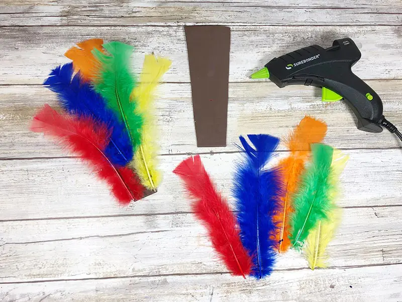 Assorted colors of craft feathers being hot glued to a piece of brown craft foam.