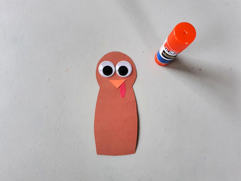 Googly eyes, beak , and snood attached to brown turkey head and neck. Assembled turkey face on light gray table next to glue stick.