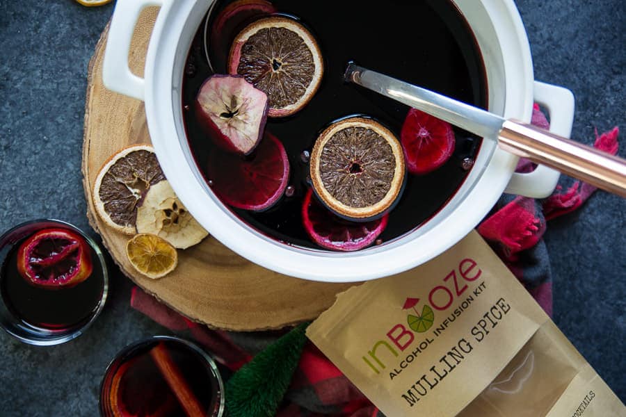 Overhead view of white punch bowl with infused mulling spice from InBooze next to glass of wine with cinnamon stick and InBooze package laying on table.