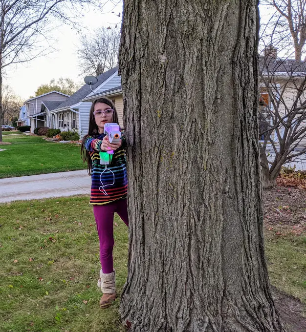 White girl wearing laser tag chest receiver and pointing Laser X Revolution blaster while standing next to tree.