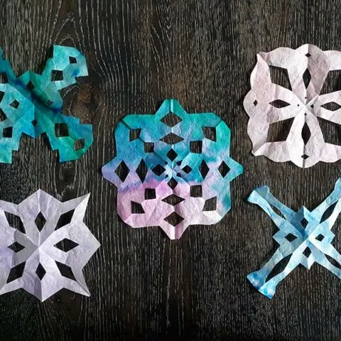 Overhead view of five finished coffee filter snowflake crafts cut in different shapes and patterns and in different colors laying on dark wooden table.