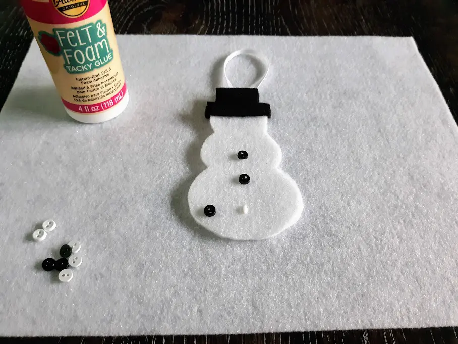 Felt snowman laying on white felt on table and gluing mini black buttons to the body.