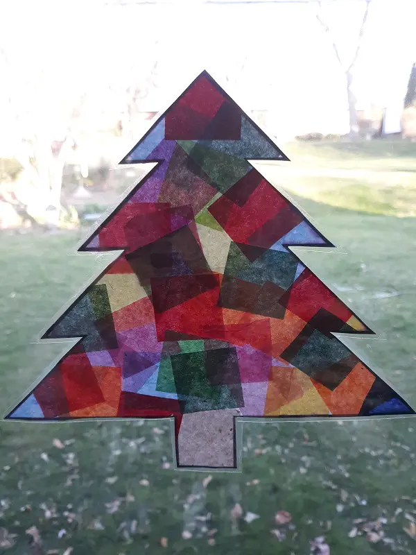 Finished evergreen tree shaped suncatcher made with tissue paper on window. Can see grass and leaves outside on the lawn.