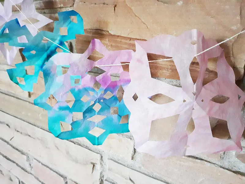 Angled view of several coffee filter snowflakes strung up as garland hanging from a brick fireplace mantel.