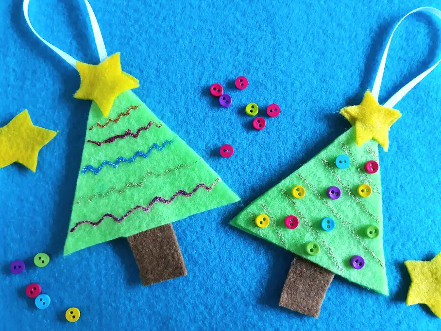 Overhead view of two Christmas tree felt ornaments. One decorated with lines of glitter glue and the other decorated with glitter glue and multicolored mini buttons. Ornaments laying on a blue felt background with stars and buttons scattered around.