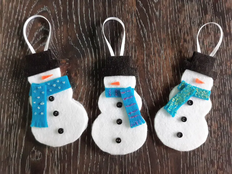 Overhead view of three completed felt snowman Christmas ornaments laying on dark wooden table.