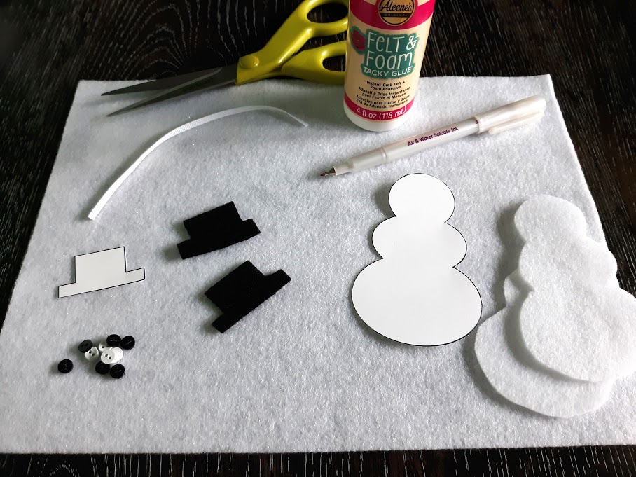 Snowman ornament pattern pieces laying on white felt along with cut out snowman bodies and hats. Other supplies also on table: pen, ribbon, glue, scissors.