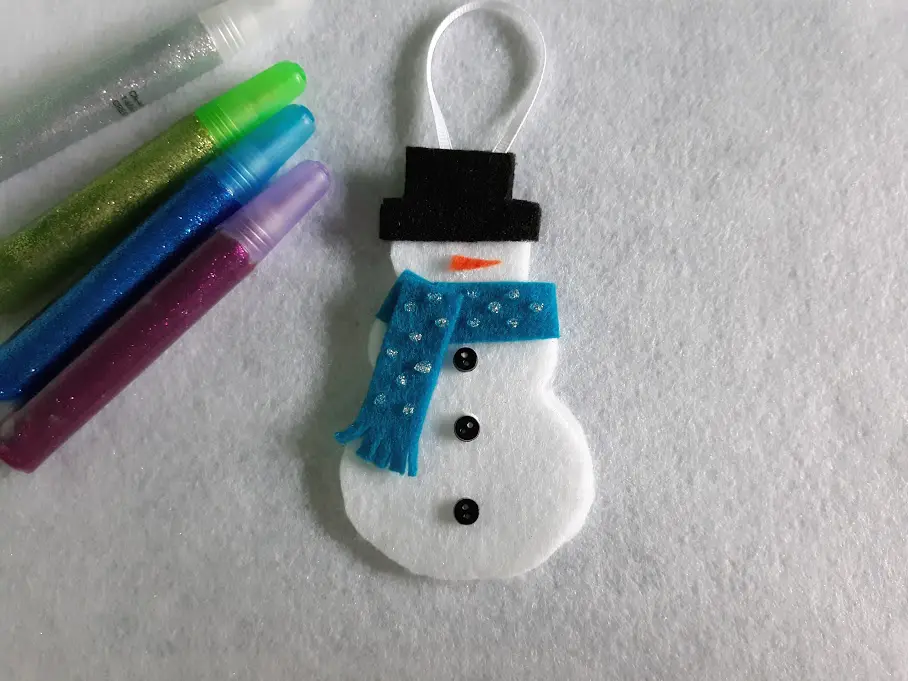 Completed felt snowman ornament laying next to glitter glue pens. Blue felt scarf on snowman decorated with dots of silver glitter glue.