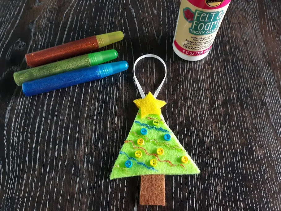 Decorated Christmas Tree ornament made out of felt laying on dark wooden table next to glitter glue pens and felt and foam tacky glue.