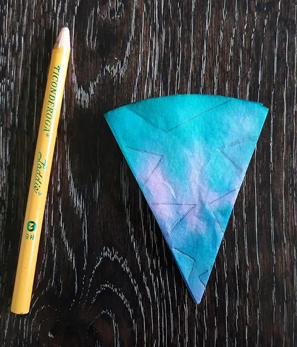 Pencil laying on dark wooden table next to blue, green, and pink tie dyed coffee filter folded like a triangle with lines drawn on it to show where to cut.