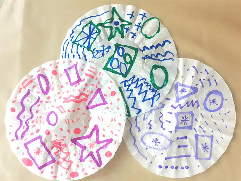 Three round coffee filter with a variety of designs drawn on them with markers laying overlapped on piece of parchment paper.