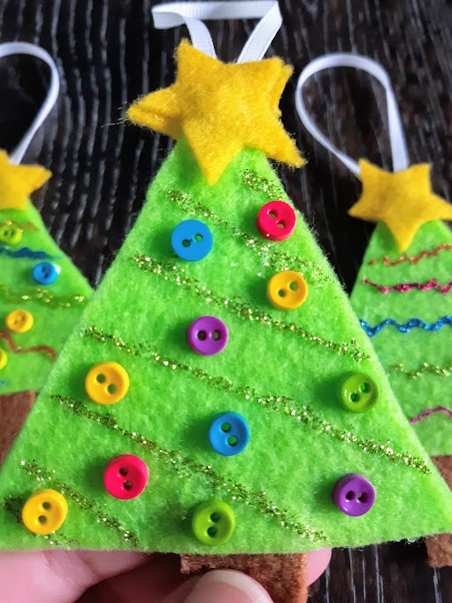 Close up view of white person's hand holding up finished felt Christmas tree ornament decorated with glitter glue and buttons. Two more finished projects laying on table in background.