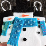 Close up of white person's hand holding a completed felt snowman ornament with two more felt snowmen laying on table in background.