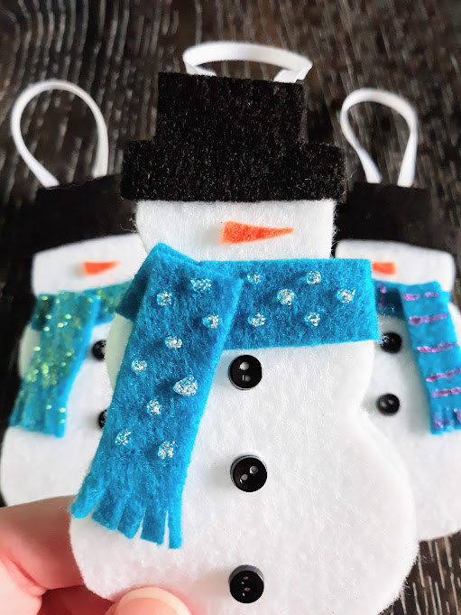 Close up of white person's hand holding a completed felt snowman ornament with two more felt snowmen laying on table in background.