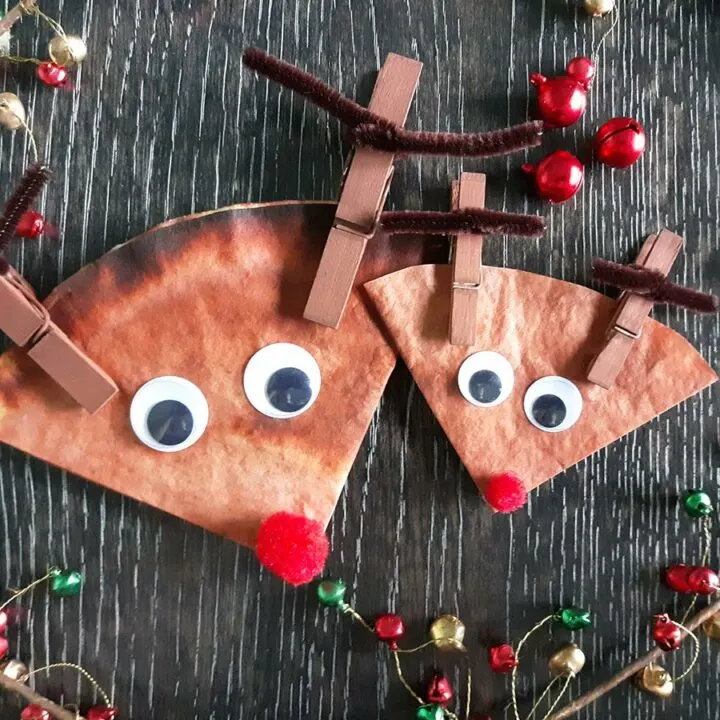 Overhead view of big and small coffee filter reindeer next to each other on dark background surrounded by small red, green, and gold decorations.