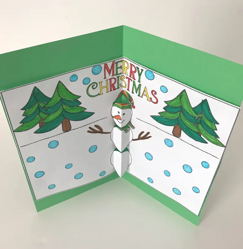 Top down view of finished snowman pop up card standing up on white table. Snowman page is colored with marker and glued to green cardstock paper.