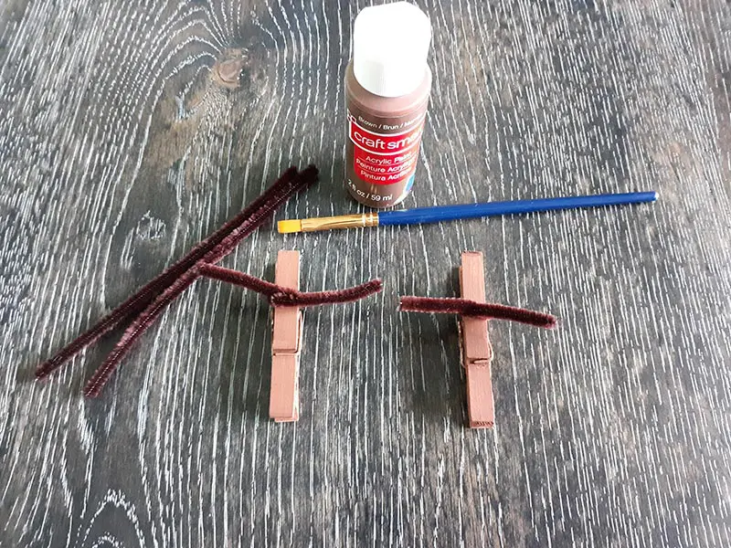 Two clothespins painted brown with brown chenille stems twisted around to make antlers. Also on table is bottle of brown paint, paint brush, and additional brown pipe cleaners.