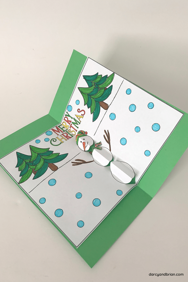 Finished pop up snowman card colored with markers and glued to green cardstock laying open at an angle on white background.