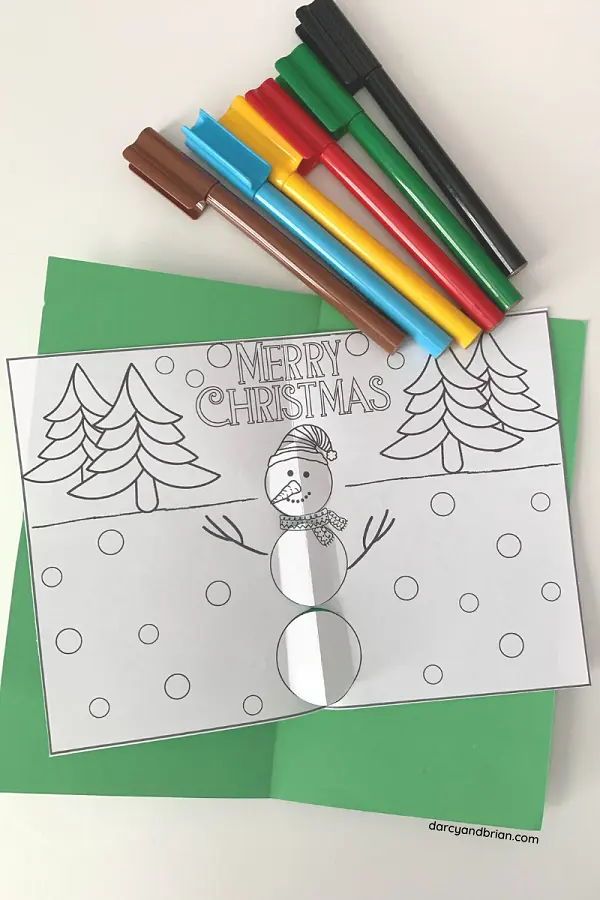 Overhead view of markers laying on white table next to green cardstock and snowman page cut and folded to create pop up effect.