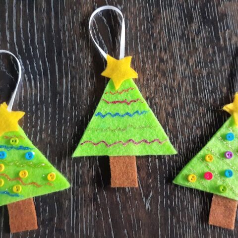 Three complete felt Christmas tree ornaments laying in a row on a dark wooden table.