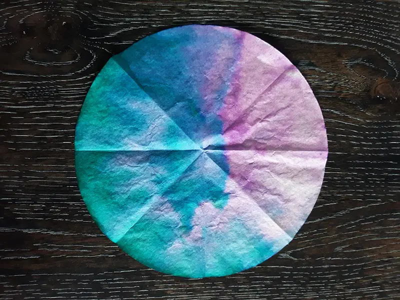 Round coffee filter tie dyed blue, green, and pink, laying on dark wooden table.