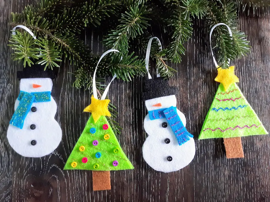 Four completed felt ornaments, alternating snowmen and Christmas trees. Ribbon is looped over evergreen branches. Background is a dark wood.