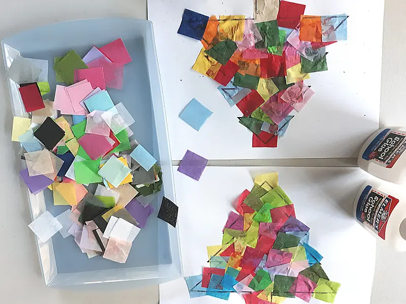 Light blue tray with assorted colors of pre-cut tissue paper squares next to two Christmas tree suncatcher templates covered with glued on tissue paper and two bottles of school glue.