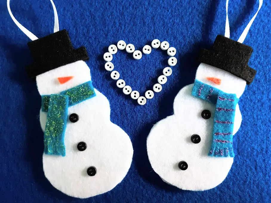 Two finished felt snowman ornaments laying on blue felt background with white mini buttons arranged in a heart between the two snowmen.
