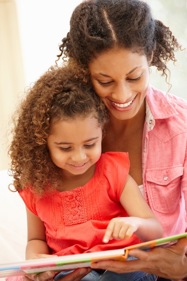 Black woman reading with daughter sitting in her lap, both smiling as little girl points at the page.