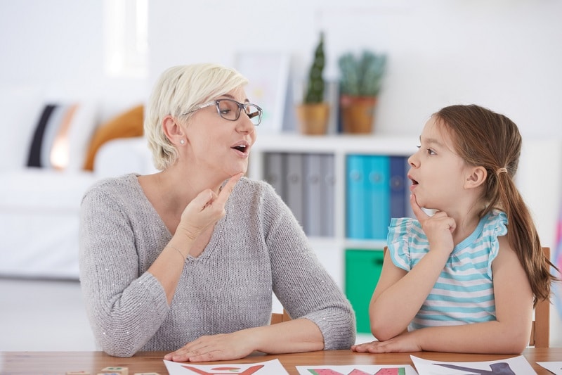 Older white lady with short hair and glasses touching chin while saying letter sound. Young white girl with dark hair in ponytail copying what teacher is doing.