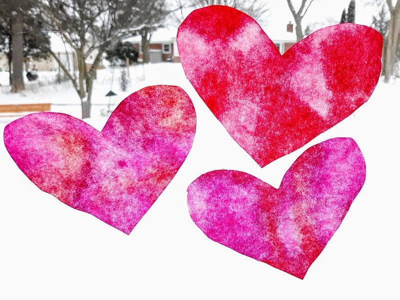 Three pink and red coffee filter hearts in a window with snow outside behind them.