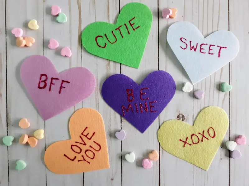 Six different colored felt hearts (pink, orange, purple, green, yellow, and white) with words written in glitter glue (BFF, Love You, Cutie, Be Mine, XOXO, Sweet) with candy conversation hearts spread out around them.