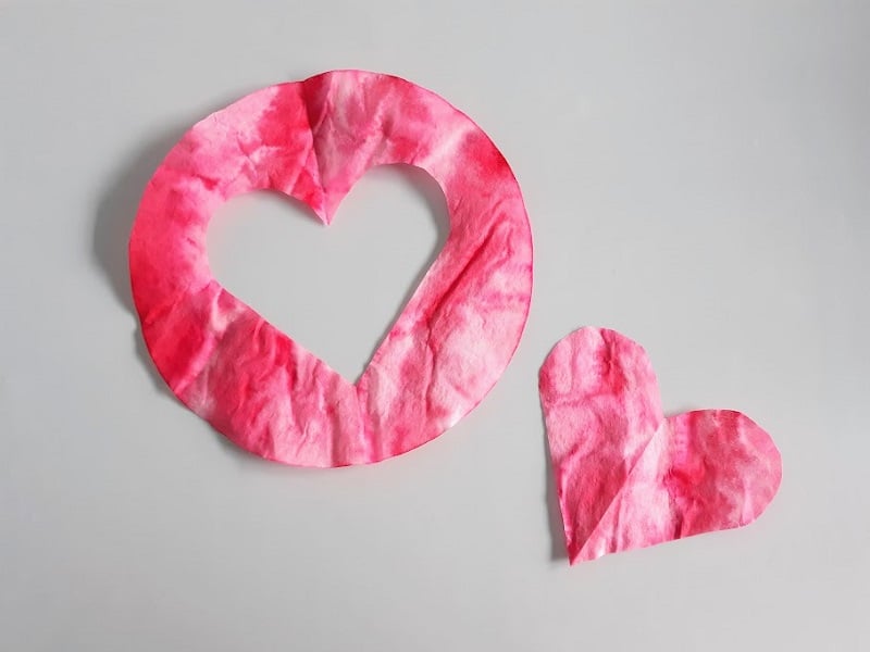 Red and pink dyed coffee filter with heart shape cut out of the center laying next to the heart cut out.
