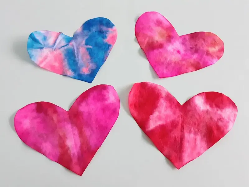 Four completed coffee filter hearts laying on an off white craft mat. One heart is blue and pink tie dyed and the other three are various shades of red and pink.