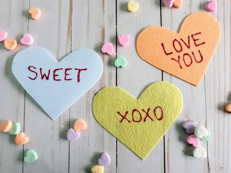 Close overhead view of three felt conversation hearts. White heart says Sweet, yellow heart says XOXO, orange heart says Love You. Small candy hearts sprinkled around the felt hearts on a light white wood background.