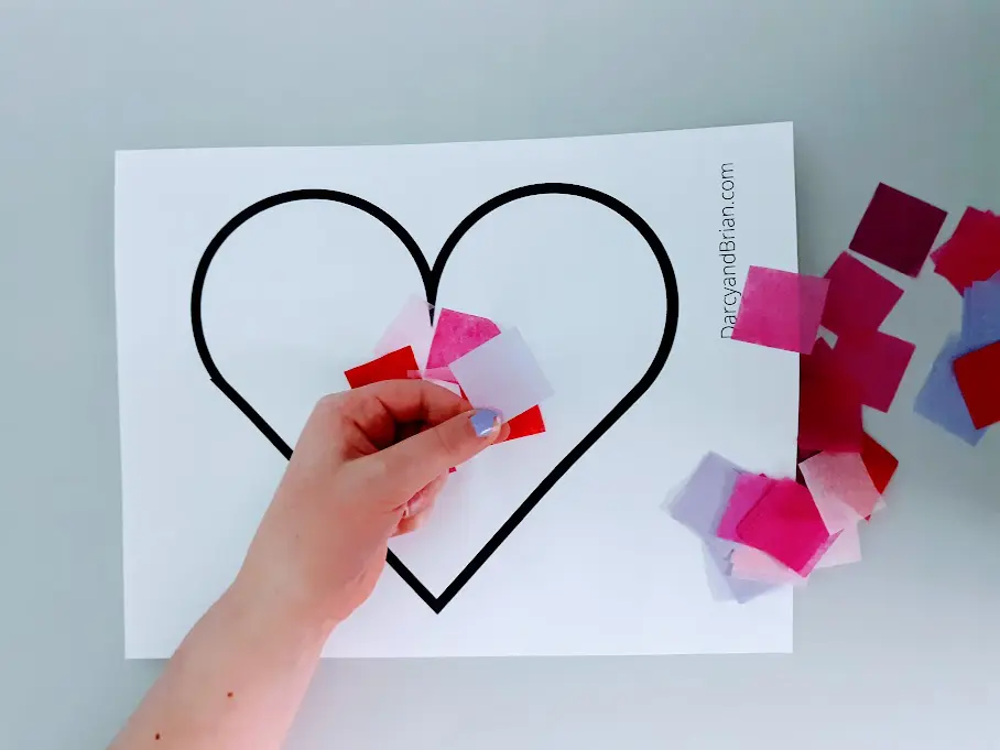 Overhead view of a young white girl's hand placing pre-cut squares of tissue paper onto heart craft template.