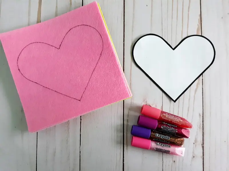 Heart pattern traced on pink felt and next to assorted glitter glue colors.