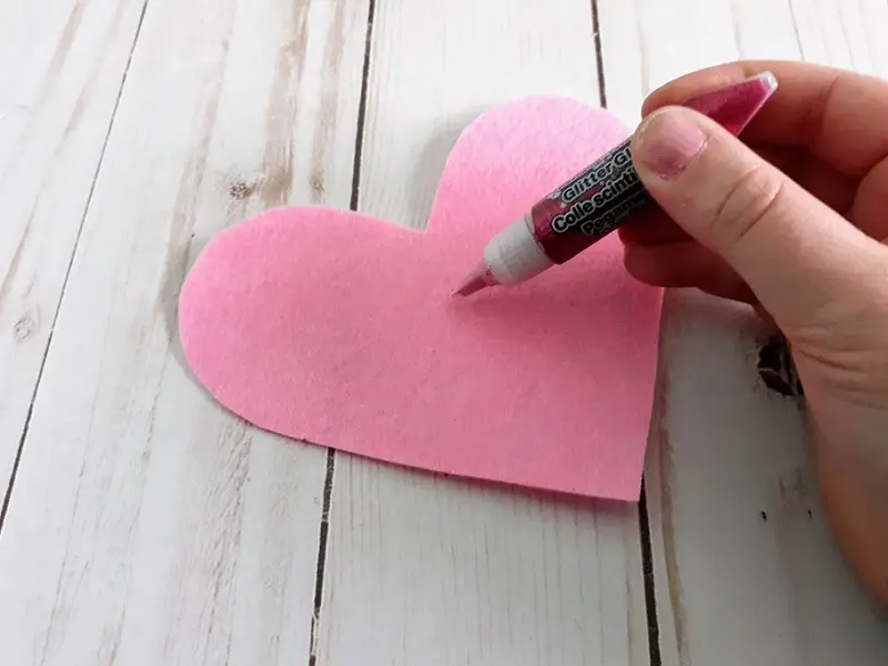 White girl's hand holding glitter glue getting ready to write and decorate pink felt heart.