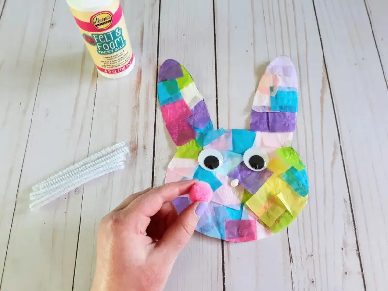 White girl's hand gluing pink pom pom nose to tissue paper bunny head. Bunny also has two wiggly eyes. White pipe cleaner pieces lay next to bunny head along with glue.