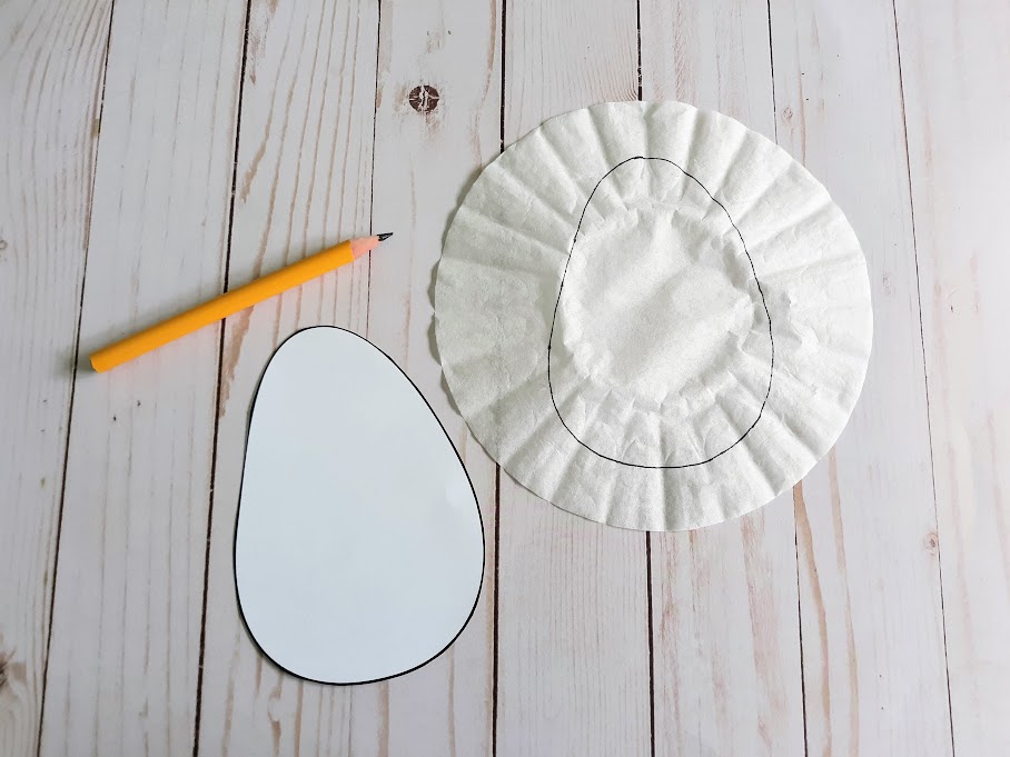 Cut out egg craft template next to a pencil and a coffee filter with traced egg shape drawn on it.