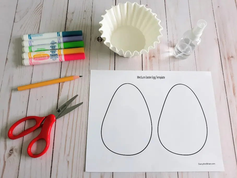 Overhead view of craft supplies to make coffee filter Easter eggs: Scissors, pencil, washable markers, coffee filters, spray bottle, and egg craft template printable.