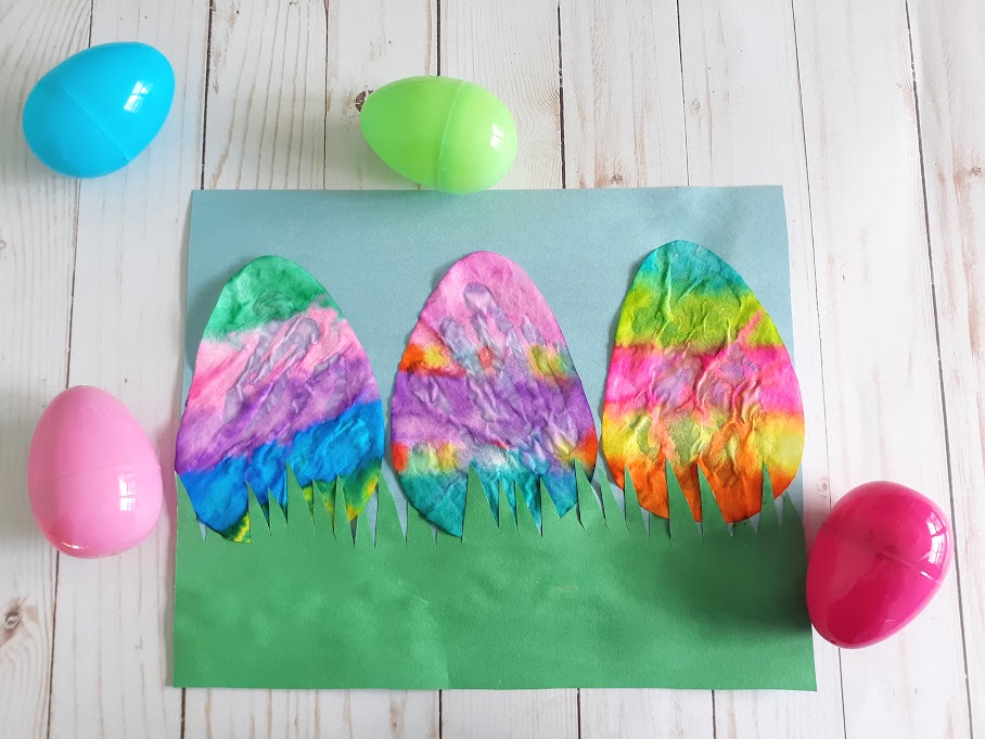 Overhead view of three finished coffee filter Easter eggs glued to blue construction paper with green fringe cut construction paper along the bottom. Four plastic eggs laying around the craft project.